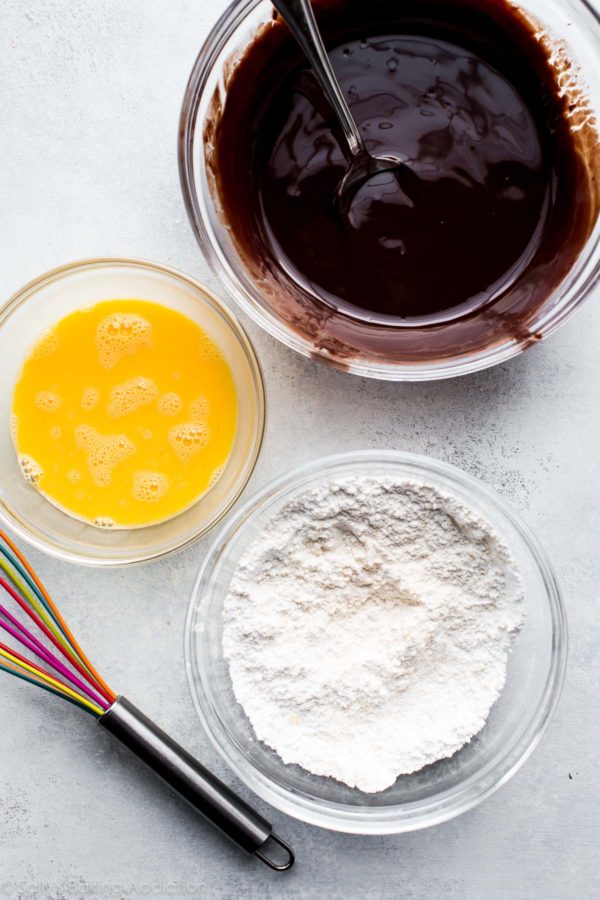 a desk with a bowl of melted chocolate, a bowl of whisked eggs and a bowl of flour with a whisk, preparation of lava cakes